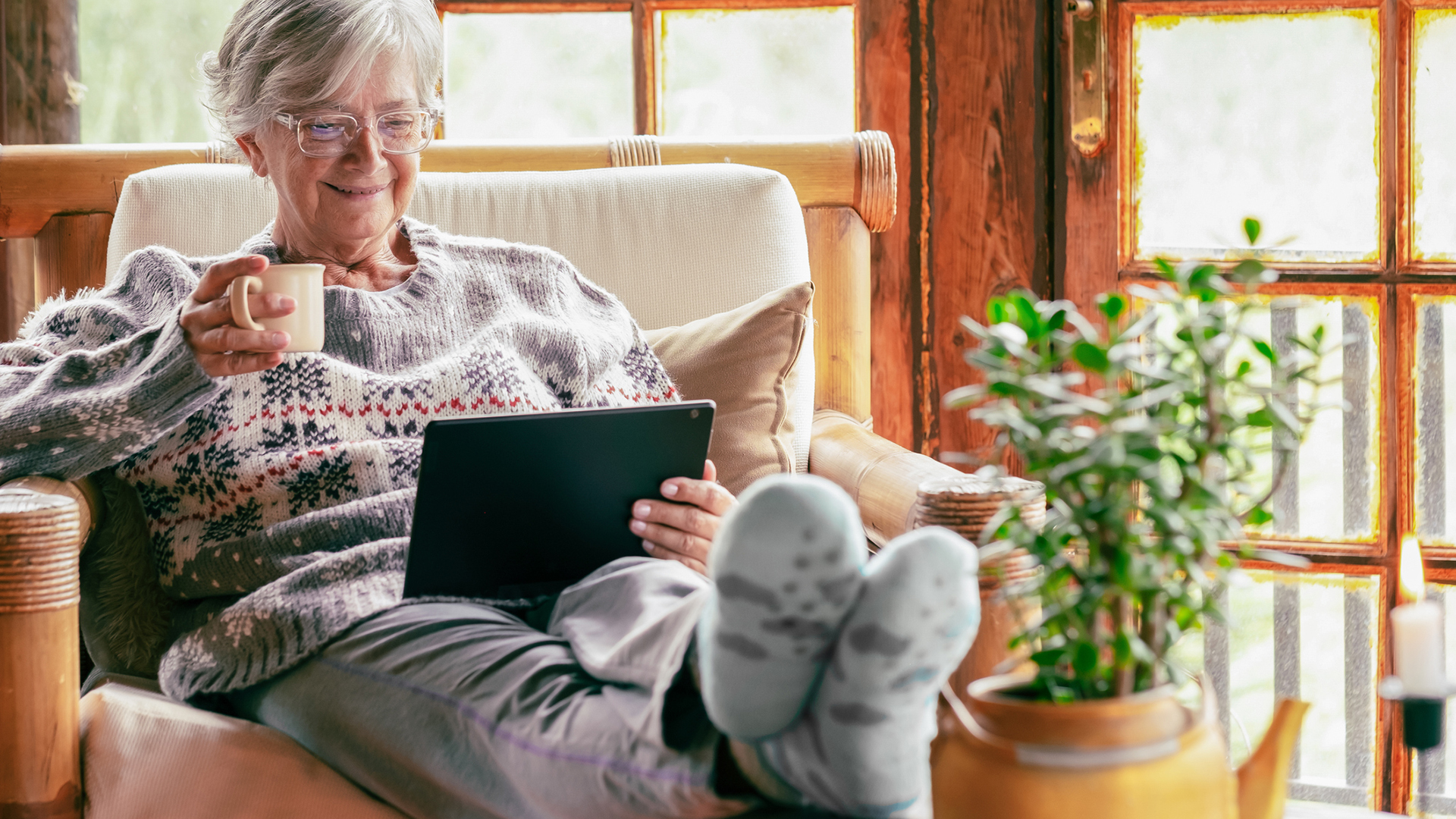 senior woman sitting at home on armchair using digital tablet wearing a warm sweater and eyeglasses