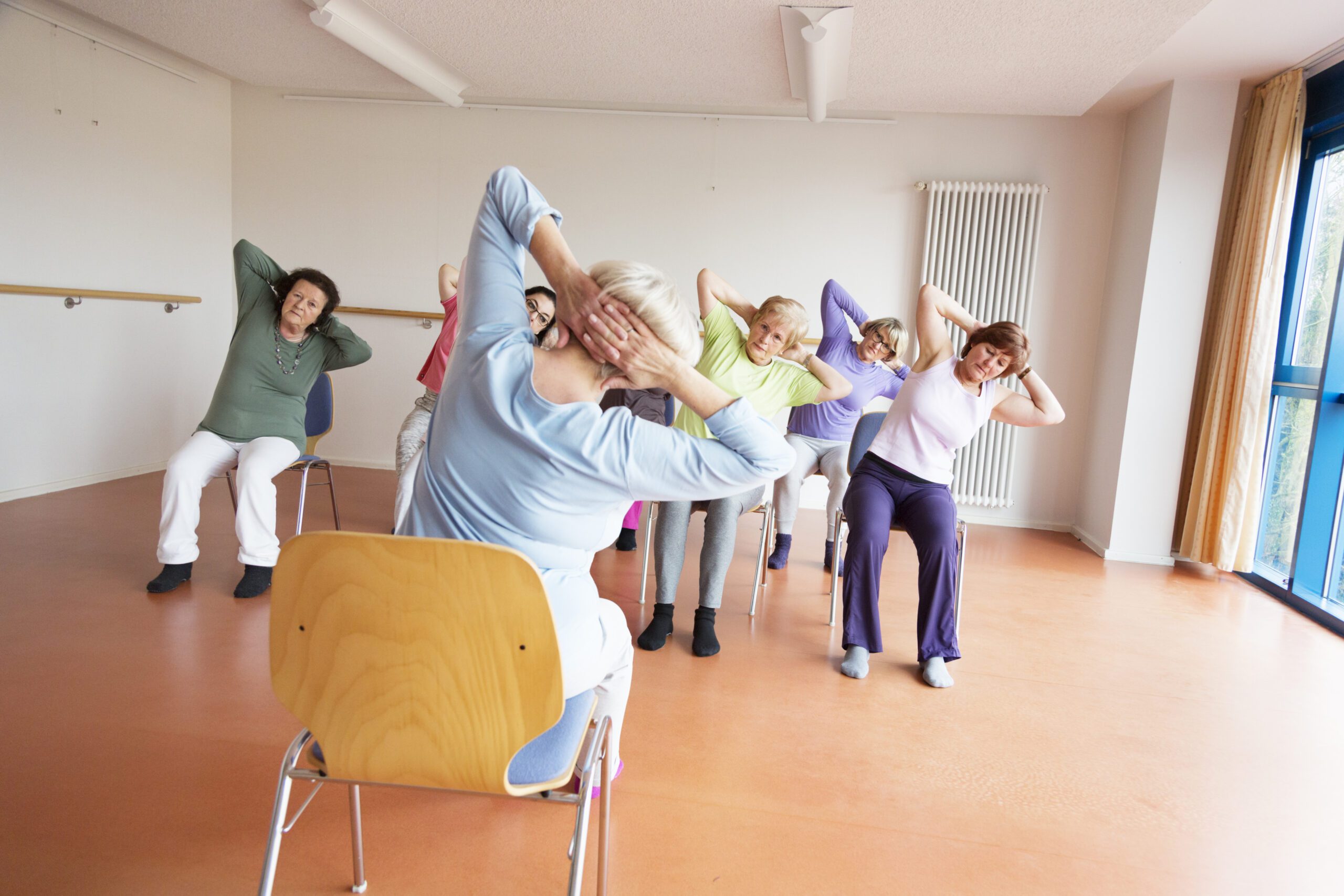 senior women exercising yoga and pilates sitting on chairs, following the instruction of their teacher